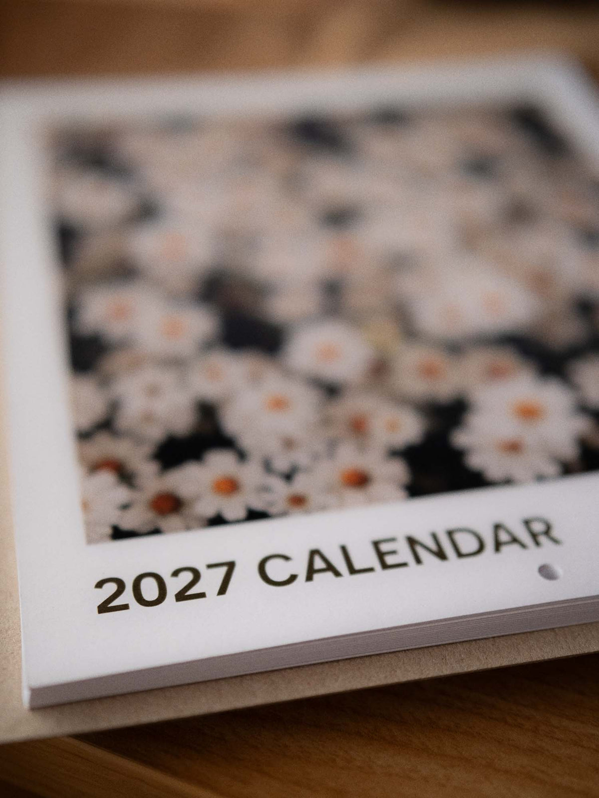 A 2027 Emerden wall calendar featuring a close-up floral photograph of white daisies with orange centers (zinnia - white star) on a dark background, shown resting on a wooden surface.